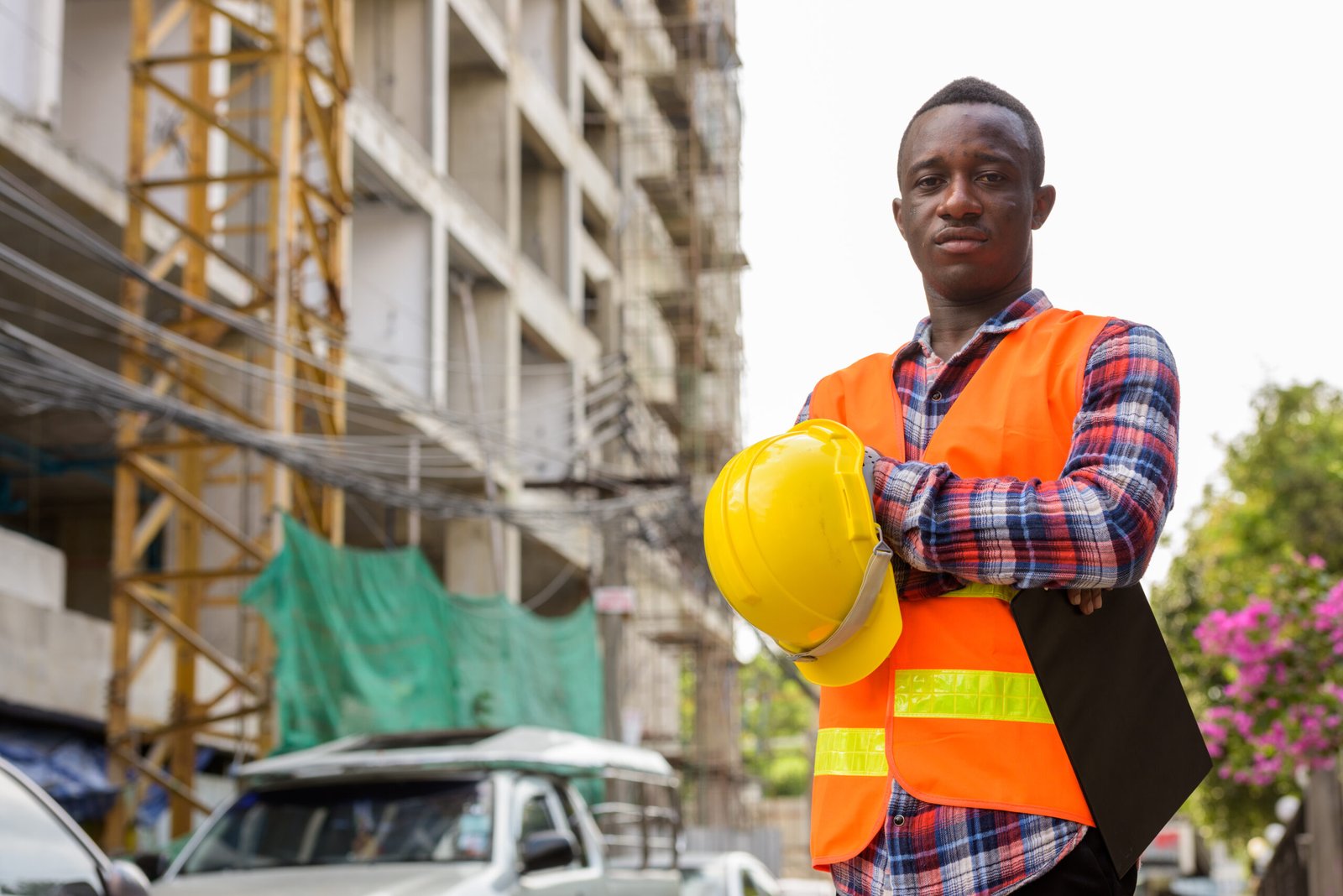 Young black African man construction worker holding clipboard and hard hat with arms crossed at building site